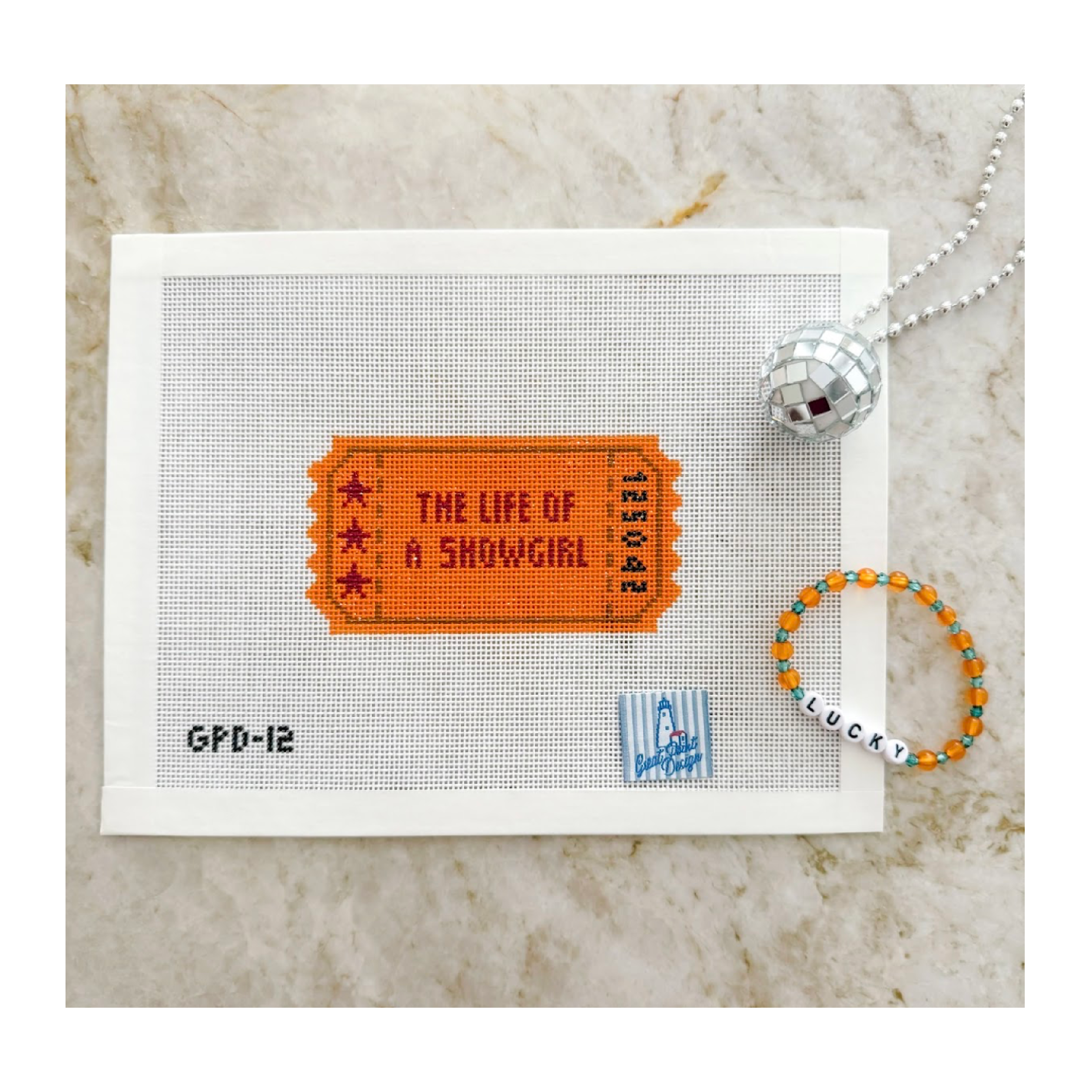 Needlepoint ticket design on fabric with a disco ball and bracelet on a marble surface